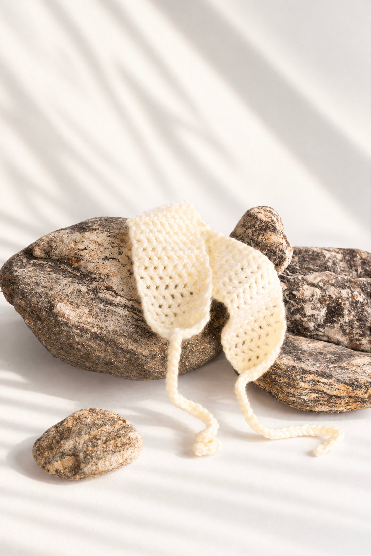 The ivory crochet headband resting over a river stone in warm afternoon light, its two ties curling onto the cloth