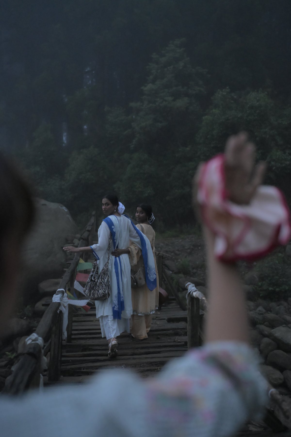 A hand lifted into the foreground holding the round, two Creatures walking a wooden bridge in the fog beyond