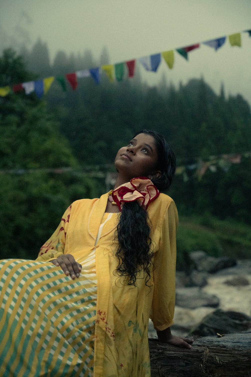 A Creature in yellow resting by a mountain river under prayer flags, the ruffled round tied in her hair